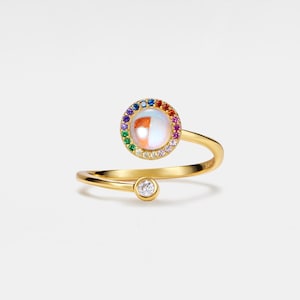 Könnte beinhalten: Ein goldener Ring mit einem Regenbogen aus bunten Steinen, die einen weißen, opalartigen Stein umgeben. Ein einzelner klarer Stein befindet sich auf der Ringschiene.