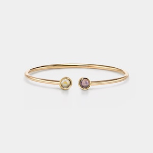 Puede incluir: Un brazalete de oro con dos piedras redondas. Una piedra es blanca con un centro amarillo, y la otra es morada.
