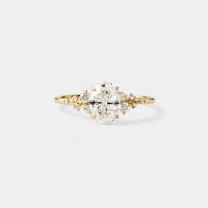 May include: A gold ring featuring a large oval-cut diamond as the centerpiece, surrounded by smaller accent diamonds. The band is a delicate gold, and the ring is set against a white background.