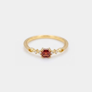 May include: A gold ring with a square, reddish-brown gemstone at the center. The band features decorative detailing with small, clear accent stones on either side of the center stone. The ring is set against a white background.