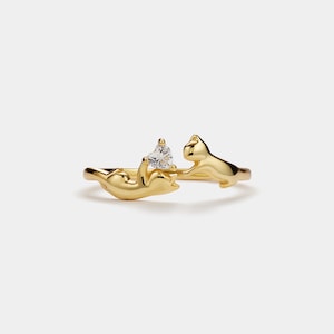 May include: A gold-colored ring featuring two stylized cat figures. One cat appears to be holding a heart-shaped, clear gemstone. The ring is designed with a minimalist aesthetic, suitable for everyday wear.
