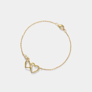 May include: A delicate gold bracelet featuring two intertwined heart pendants. The bracelet has a fine chain and a lobster clasp closure. The hearts are open-worked and connected, symbolizing love and connection. A small cluster of crystals adds a touch of sparkle.