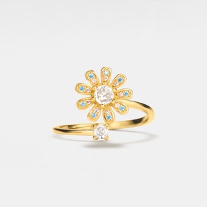 May include: A gold ring with a delicate daisy design featuring a clear center stone and smaller blue and pink stones around the edges. The ring is open and wraps around the finger.