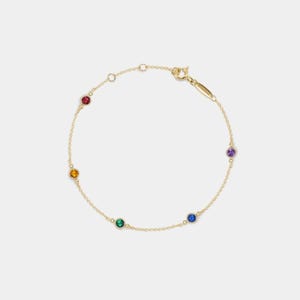 Puede incluir: Un brazalete de cadena dorada con cinco pequeñas piedras preciosas en los colores del arcoíris: rojo, naranja, amarillo, verde y morado.