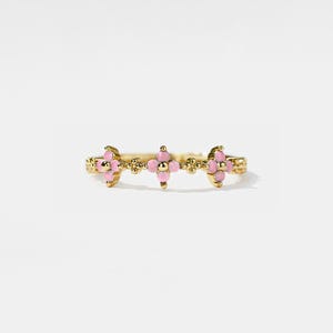 Puede incluir: Un anillo dorado con un diseño floral y piedras preciosas rosas. El anillo tiene una apariencia delicada y ornamentada con tres grupos de piedras rosas en forma de flor. El anillo está sobre un fondo blanco.
