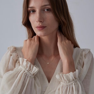 May include: A woman wearing a white blouse with ruffles on the sleeves. She is wearing a delicate gold necklace with a small pendant.