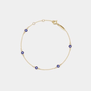 Puede incluir: Una delicada pulsera de cadena dorada con cinco dijes circulares espaciados uniformemente. Cada dije muestra un motivo de ojo turco en azul, blanco y negro. La pulsera incluye un pequeño cierre dorado y una fina etiqueta metálica cerca del cierre.