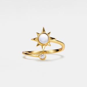 May include: A gold ring with a moonstone sun design and a small clear stone accent. The ring is open and adjustable.