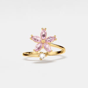May include: A gold ring with a pink flower design and a single clear stone accent. The ring is a delicate and feminine piece of jewelry.