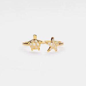 Puede incluir: Anillo de oro con un diseño de tortuga y estrella de mar, ambos adornados con piedras de circonita cúbica transparentes.