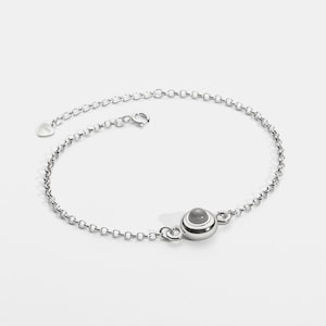 May include: A silver anklet with a small, round, gray stone pendant. The anklet is made of a delicate chain with a lobster clasp closure. The pendant is accented with white and silver bands. The anklet is displayed on a white background.