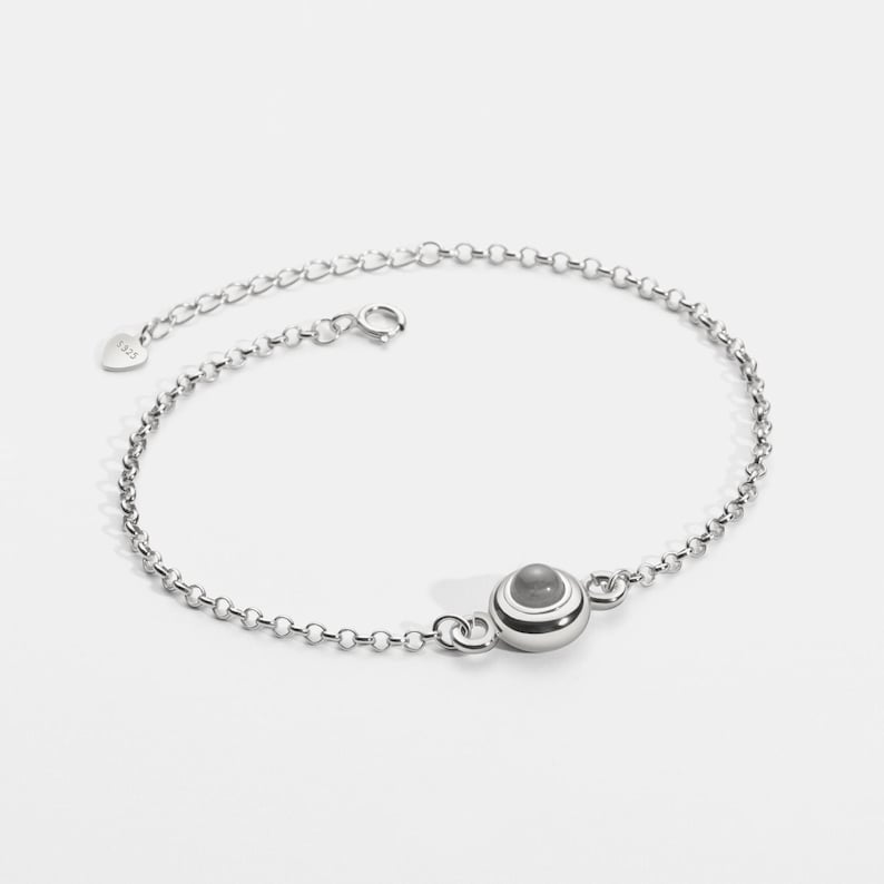 May include: A silver anklet with a small, round, gray stone pendant. The anklet is made of a delicate chain with a lobster clasp closure. The pendant is accented with white and silver bands. The anklet is displayed on a white background.