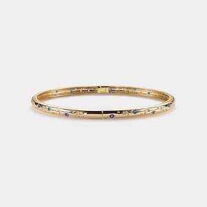 May include: A gold bangle bracelet with a hinged clasp. The bracelet is decorated with small blue and clear gemstones in a floral pattern. The bracelet is approximately 6.35 cm in diameter.