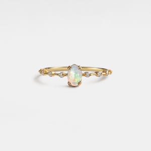 Puede incluir: Un delicado anillo de oro con una piedra preciosa de ópalo ovalada como pieza central. La banda está adornada con pequeñas piedras preciosas redondas y transparentes. El ópalo muestra un juego de colores, incluyendo blanco, rosa y verde. El anillo está sobre un fondo blanco.