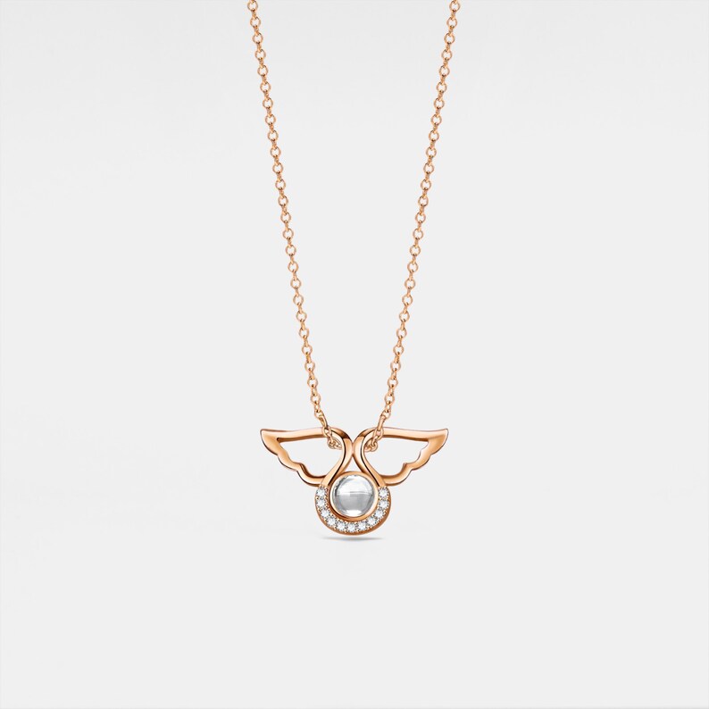 May include: A rose gold necklace with a delicate angel wing pendant featuring a small round gemstone surrounded by smaller diamonds.