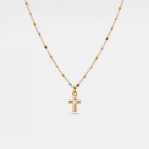 Puede incluir: Collar dorado con un colgante de cruz con una gema transparente en el centro. La cadena está adornada con pequeñas cuentas de colores en tonos amarillo, azul, marrón y morado. Una joya delicada y elegante.