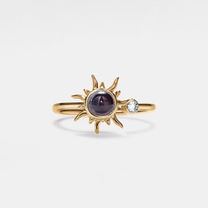 May include: Gold ring with a sun design featuring a dark purple gemstone and a small clear gemstone.