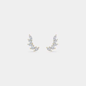 May include: A pair of gold-tone earrings, each with a curved arrangement of clear, marquise-cut gemstones. The earrings are displayed against a plain, light backdrop, emphasising their delicate and elegant design. The gemstones are arranged to resemble a leaf or feather.