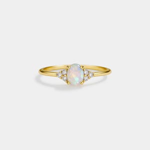 May include: A gold ring featuring an oval opal gemstone, flanked by clusters of small, round, clear gemstones. The opal has a pearlescent, iridescent quality. The ring band is thin and gold-colored, set against a white background.