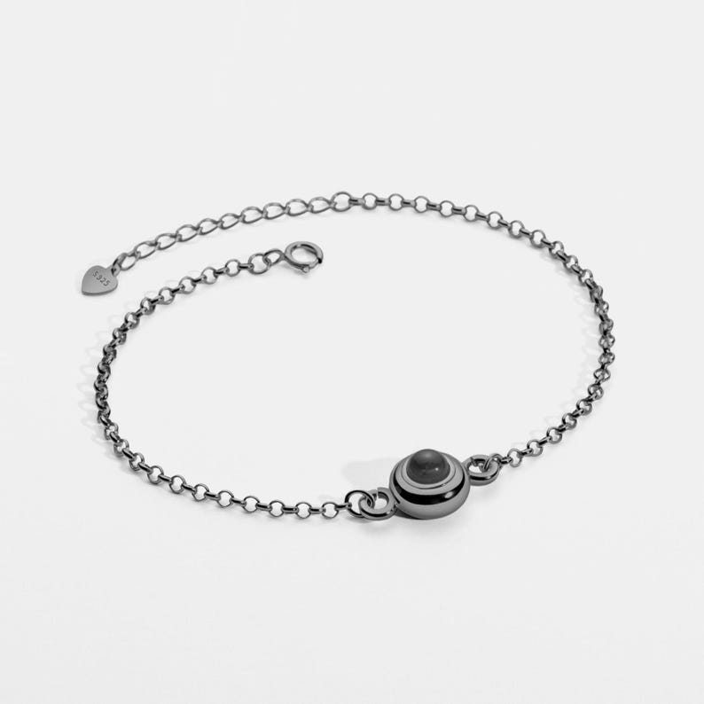 May include: A delicate silver bracelet with a small, round, gray and white stone. The bracelet features a chain design with a small heart-shaped tag near the clasp. The bracelet is shown on a white background.