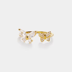 Könnte beinhalten: Ein goldfarbener Ring mit zwei Schmetterlingsmotiven. Einer ist mit klaren, marquiseförmigen Steinen besetzt, der andere mit kleineren, runden, klaren Steinen. Der Ring hat ein zartes, elegantes Design, passend für jeden Anlass.