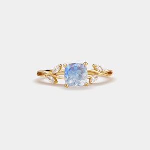 Puede incluir: Un anillo de oro con una gran piedra preciosa azul iridiscente de forma cuadrada. La banda del anillo tiene un diseño en forma de hoja con pequeñas piedras de acento transparentes. El anillo está sobre un fondo blanco liso.