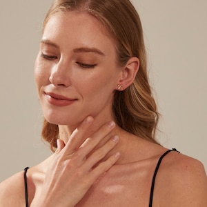 May include: A woman with long blonde hair wearing a black vest top and small gold earrings. She is touching her neck with her hand.