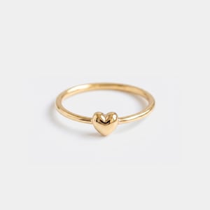 May include: A gold-colored ring with a heart-shaped charm. The ring band is thin and smooth, and the heart charm is centered on the band. The ring is set against a white background.