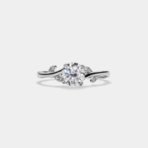 May include: A silver engagement ring featuring a round, clear diamond set in a delicate, vine-like band. The band is adorned with small, sparkling diamonds that resemble leaves.
