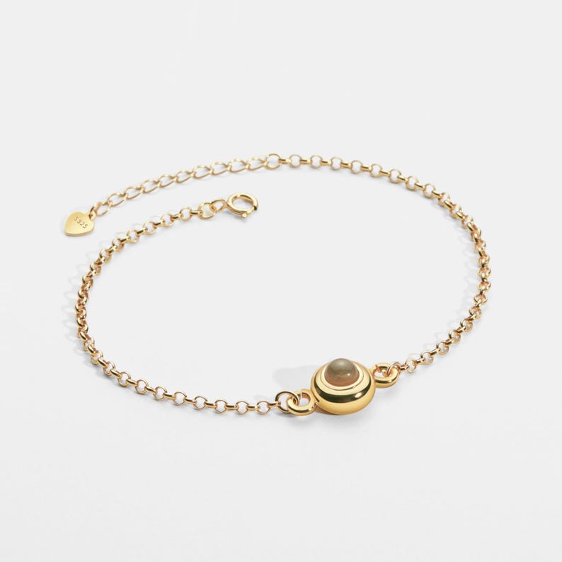 May include: A delicate gold-colored bracelet featuring a small, round, light-colored stone set in a gold bezel. The bracelet has a delicate chain design with a lobster clasp closure. The metal is stamped with "S925".