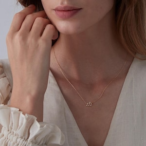 May include: A delicate gold necklace with a small, minimalist pendant featuring three curved lines. The necklace is worn by a person with a light complexion and long brown hair.