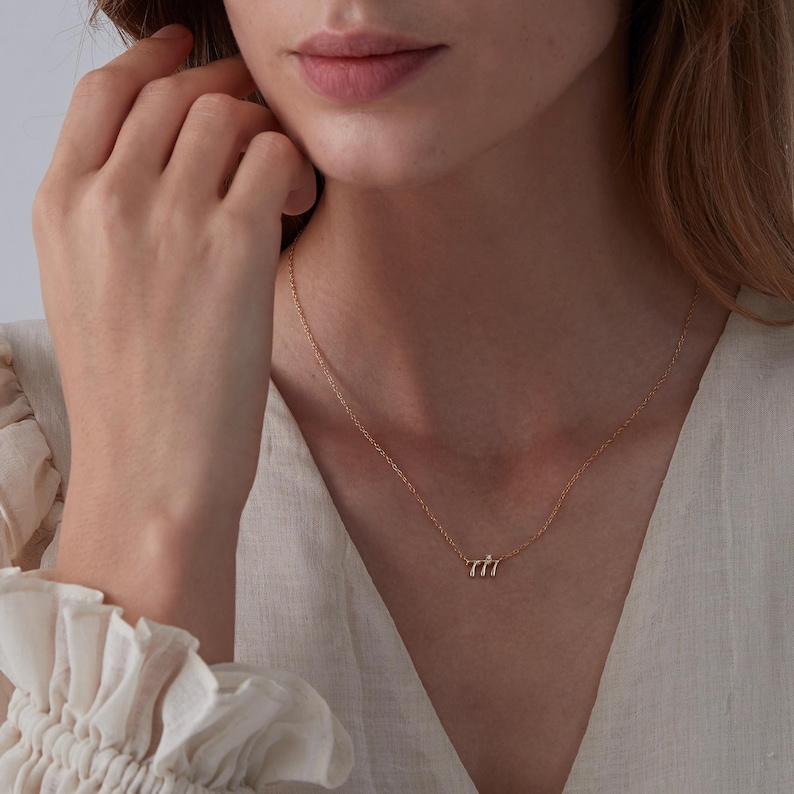 May include: A delicate gold necklace with a small, minimalist pendant featuring three curved lines. The necklace is worn by a person with a light complexion and long brown hair.