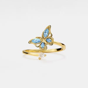 Könnte beinhalten: Ein goldener Ring mit einem blauen Schmetterlingsdesign. Der Schmetterling hat klare Kristall-Akzente. Der Ring ist offen und verstellbar.