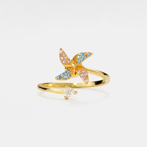 May include: A gold ring with a colorful pinwheel design and a single clear stone. The pinwheel has blue, pink, and yellow accents.