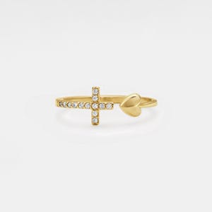 May include: A gold-colored ring featuring a cross design adorned with small, clear gemstones and a heart-shaped accent. The ring is set against a plain white background, highlighting its delicate details and craftsmanship.