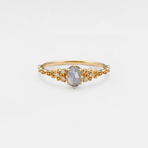 Può includere: Un delicato anello d'oro con una pietra ovale grigia circondata da piccoli diamanti. La fascia presenta un design floreale.