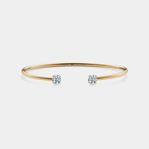 May include: A gold-colored open cuff bracelet with two round, clear gemstones set at each end. The bracelet has a simple, elegant design, and the gemstones sparkle against the gold. The bracelet is shown on a white background.