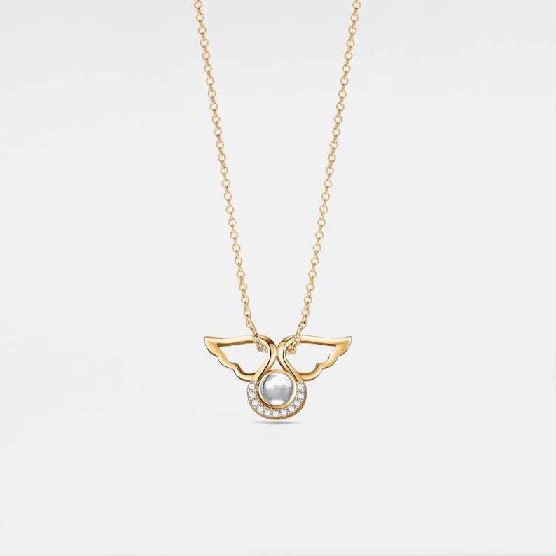 May include: A gold necklace with a pendant featuring a pair of angel wings with a small round gemstone in the center. The gemstone is surrounded by a halo of smaller gemstones.