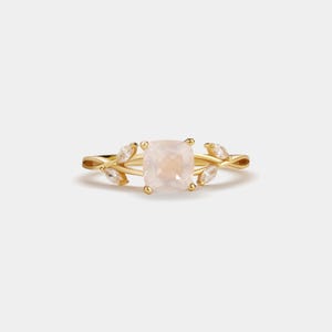 May include: A gold ring featuring a square, light pink gemstone at the center, flanked by leaf-shaped accents. The ring's band and leaf details have a polished finish, creating a delicate and elegant design. The ring is set against a plain white background.
