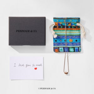 May include: A small, brown, metal pendant necklace in a blue and orange patterned drawstring bag. The bag is on a white surface. A handwritten note reads "I love you so much" with a red heart. A black box with the text "PERIMADE & CO." is also on the surface.