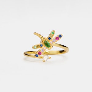 May include: A gold ring with a colorful dragonfly design. The dragonfly has green, blue, pink, and yellow accents. The ring is a delicate piece of jewelry.