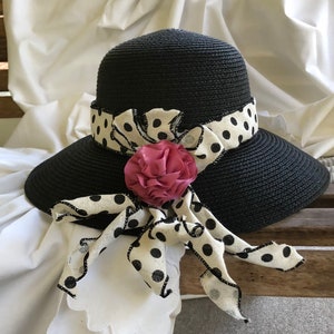 Könnte beinhalten: Ein schwarzer Strohhut mit breiter Krempe und einem weißen und schwarzen gepunkteten Band. Eine rosa Stoffblume ist am Band befestigt.