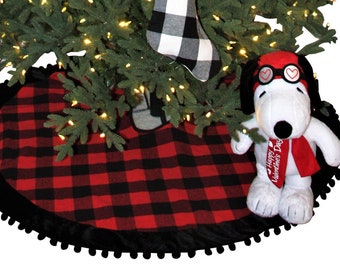 May include: A Christmas tree decorated with red and black buffalo check stockings. The tree is adorned with white lights and has a black and red buffalo check tree skirt. A white plush dog wearing a red scarf and goggles is sitting in front of the tree.