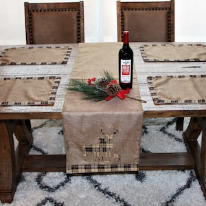 May include: A rustic wooden dining table set with four placemats and a table runner. The placemats are brown with a plaid border, and the table runner is brown with a deer design. A bottle of wine is placed on the table runner.