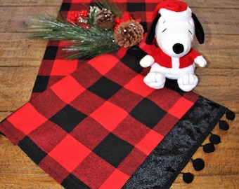 May include: Red and black buffalo plaid table runner with a black velvet trim and black pom-poms. A plush white and red Santa Claus Snoopy doll is sitting on the runner.