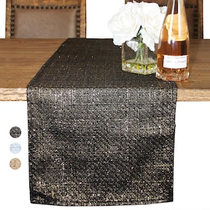 Op de afbeelding: Een zwarte en gouden glitter tafelrunner met een geweven textuur. De runner ligt op een houten tafel met een vaas met witte bloemen en een fles wijn.