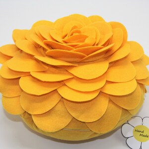 K&ouml;nnte beinhalten: Ein gro&szlig;es, rundes, gelbes Kissen aus Filz in Blumenform. Das Kissen besteht aus vielen Schichten von Filzbl&uuml;tenbl&auml;ttern, die einen 3D-Effekt erzeugen. Das Kissen hat ein Etikett mit der Aufschrift "Hand Made".