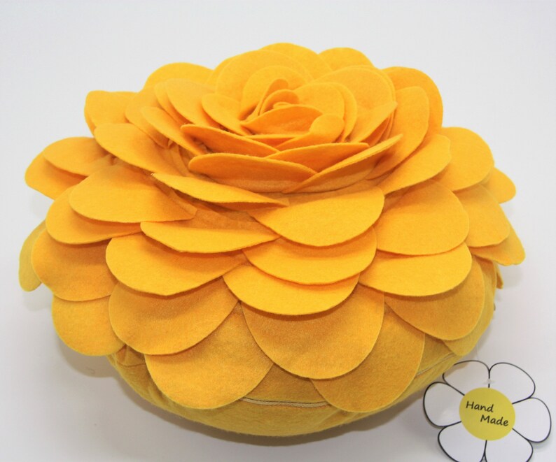K&ouml;nnte beinhalten: Ein gro&szlig;es, rundes, gelbes Kissen aus Filz in Blumenform. Das Kissen besteht aus vielen Schichten von Filzbl&uuml;tenbl&auml;ttern, die einen 3D-Effekt erzeugen. Das Kissen hat ein Etikett mit der Aufschrift "Hand Made".