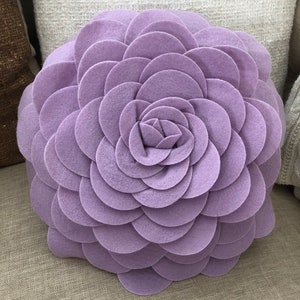 K&ouml;nnte beinhalten: Ein gro&szlig;es, rundes, lilafarbenes Kissen in Blumenform. Das Kissen ist aus weichem, pl&uuml;schem Stoff gefertigt und hat ein 3D-Design.