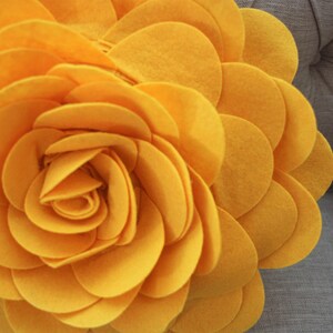 K&ouml;nnte beinhalten: Ein gro&szlig;es, gelbes Kissen in Form einer Filzblume. Die Blume besteht aus vielen Schichten von Filzkreisen, die einen 3D-Effekt erzeugen.